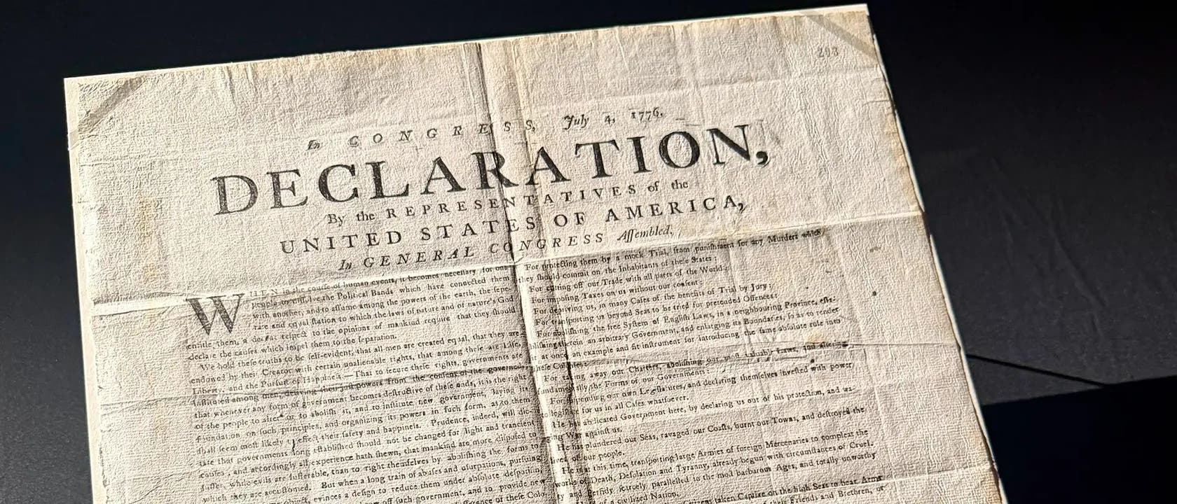 Ein Foto zeigt eine historische Kopie der Unabhängigkeitserklärung der Vereinigten Staaten von Amerika. Der Text beginnt mit dem Wort "DECLARATION" in großen Buchstaben, gefolgt von weiteren Details über die Repräsentanten und den Kongress. Das Dokument ist auf gealtertem Papier gedruckt und weist Falten und Verfärbungen auf, die auf sein Alter hinweisen. Es liegt auf einer dunklen Oberfläche, die den Kontrast zum Papier verstärkt. Ein Foto zeigt eine historische Kopie der Unabhängigkeitserklärung der Vereinigten Staaten von Amerika. Der Text beginnt mit dem Wort "DECLARATION" in großen Buchstaben, gefolgt von weiteren Details über die Repräsentanten und den Kongress. Das Dokument ist auf gealtertem Papier gedruckt und weist Falten und Verfärbungen auf, die auf sein Alter hinweisen. Es liegt auf einer dunklen Oberfläche, die den Kontrast zum Papier verstärkt.