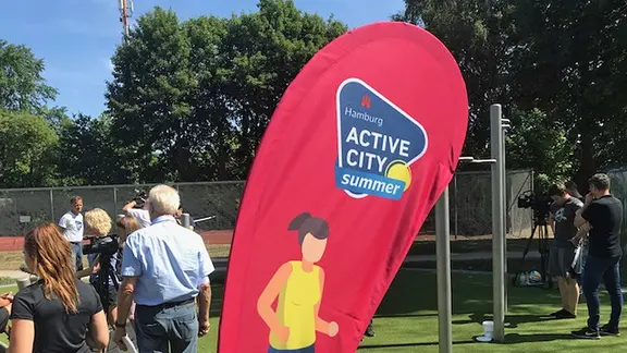 Eine rote Fahne mit der Aufschrift "Active City Summer Hamburg" steht in einem Park. | NDR Eine rote Fahne mit der Aufschrift "Active City Summer Hamburg" steht in einem Park.