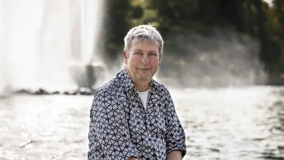 Der bisherige Intendant des "Kleinen Fests im Großen Garten" in Hannover, Casper de Vries, tritt zurück. | Landeshauptstadt Hannover / Helge Krückeberg, Helge Krückeberg Ein Mann im gemusterten Hemd sitzt vor einem Brunnen.
