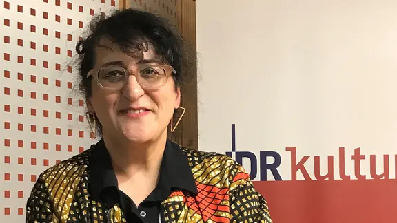 Eine Frau mit schwarzen lockigen Haaren, zum Zopf frisiert, blickt in die Kamera. Sie trägt eine Brille und ein buntes Oberteil. | NDR Eine Frau mit schwarzen lockigen Haaren, zum Zopf frisiert, blickt in die Kamera. Sie trägt eine Brille und ein buntes Oberteil.