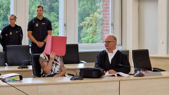 Eine Frau hält sich einen roten Hefter vor das Gesicht, links von ihr sitzt ein Mann. Im Hintergrund stehen zwei Polizist:innen.