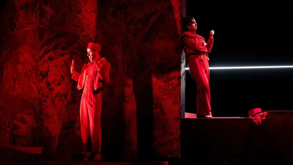 Auf einer rot beleuchteten Bühne stehen zwei Männer in roten Anzügen und lehnen sich an eine Art Felsen.