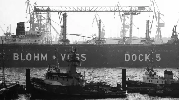 Schlepper vor dem Dock 5 der Werft von Blohm und Voss im Hamburger Hafen am 02.11.1978. | picture alliance / Klaus Rose | Klaus Rose, picture alliance / Klaus Rose | Klaus Rose Ein altes Bild vom Hamburger Hafen zeigt die Werft von Blohm + Voss und Schiffe davor.
