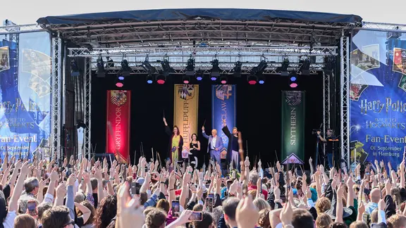 Mehrere Schaupieler und Bürgermeister peter Tschentscher stehen beim Harry-Potter-Fan-Event "Back to Hogwarts" am Theater am Großmarkt auf der Bühne. Die Zuschauer im Vorgergrund halten Zauberstäbe in die Luft. | picture alliance / dpa, Georg Wendt Mehrere Schaupieler und Bürgermeister peter Tschentscher stehen beim Harry-Potter-Fan-Event "Back to Hogwarts" am Theater am Großmarkt auf der Bühne. Die Zuschauer im Vorgergrund halten Zauberstäbe in die Luft.