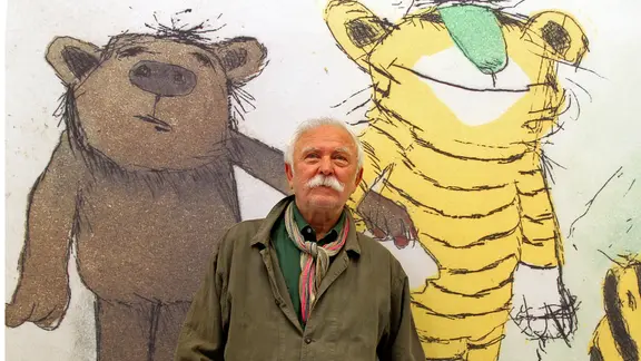 Ein Mann mit weißem Schnurrbart (Janosch) steht vor einer Zeichnung mit einem Bär und einem Tiger. | picture alliance / dpa | Roland Weihrauch Ein Mann mit weißem Schnurrbart (Janosch) steht vor einer Zeichnung mit einem Bär und einem Tiger.