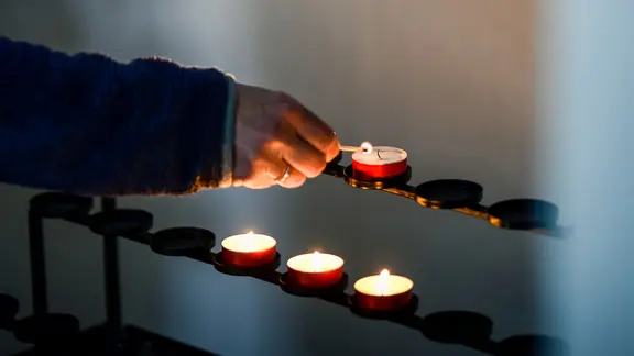 Das Bild zeigt eine Hand mit einen Streichholz, die ein Teelicht in einer Kirche anzündet.