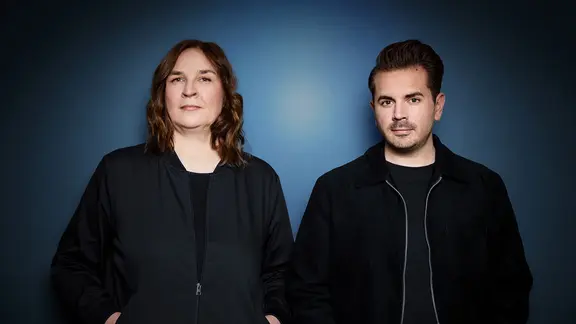 Eine Frau und ein Mann stehen vor einer blauen Wand. | Sinje Hasheider Eine Frau und ein Mann stehen vor einer blauen Wand.