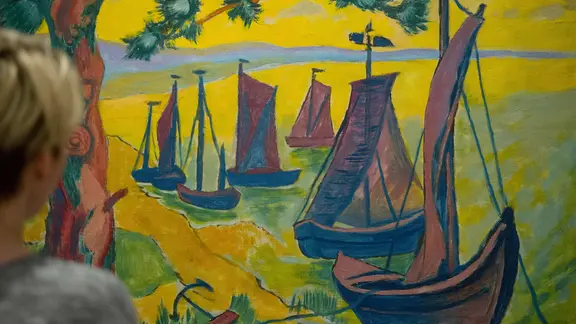 Der Künstler Max Pechstein ist vor allem für seine sogenannten Südsee-Bilder bekannt. | picture alliance/dpa/Stefan Sauer Eine Person schaut auch expressionistisches, farbintensives Bild von mehreren Segelbooten an einem Strand.
