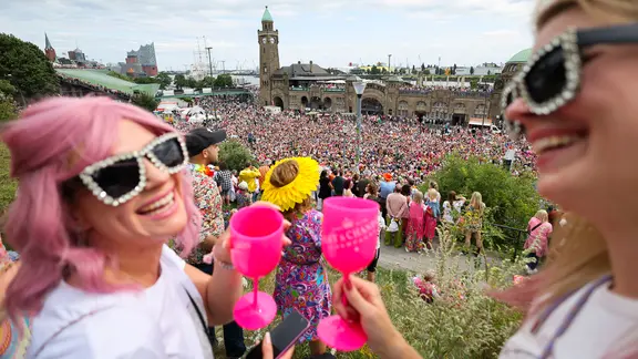 Teilnehmerinnen des 27. Schlagermoves stehen vor den Landungsbrücken und verfolgen die Schlagerkarawane durch den Stadtteil St. Pauli. Start und Ziel ist auf dem Heiligengeistfeld. | picture alliance/dpa/Christian Charisius Teilnehmerinnen des 27. Schlagermoves stehen vor den Landungsbrücken und verfolgen die Schlagerkarawane durch den Stadtteil St. Pauli. Start und Ziel ist auf dem Heiligengeistfeld.
