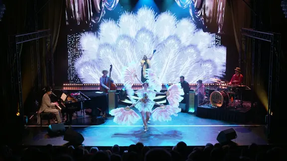 Die 20er-Jahre-Show "Glanz auf dem Vulkan" mit Tänzerinnen und riesigen Federn. | Screenshot Riesige weiße Federn schmücken das Bühnenbild. Auf der Bühne ist eine fünfköpfige Band, drei Tänzerinnen und eine Frau, die singt.