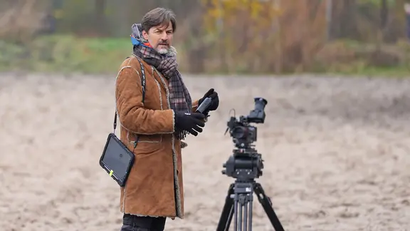 Der Regisseur Hans Steinbichler in warmer Kleidung mit Schal neben einem Stativ am Set einer Tatort-Folge in Niedersachsen