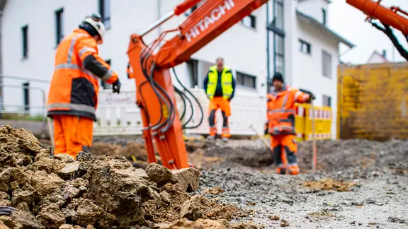 Drei Bauarbeiter auf einer Baustelle mit einem Schaufelbagger. | dpa-Bildfunk, David Inderlied Drei Bauarbeiter auf einer Baustelle mit einem Schaufelbagger.