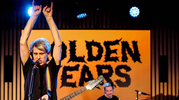 Feiern in Dresden beim Open Air im Tante Ju ihre "Golden Years": Tocotronic | IMAGO / Votos-Roland Owsnitzki Dirk von Lowtzow mit Gitarre auf der Bühne, die Arme in die Luft gerissen. Im Hintergrund ein Banner mit der Aufschrift Golden Years