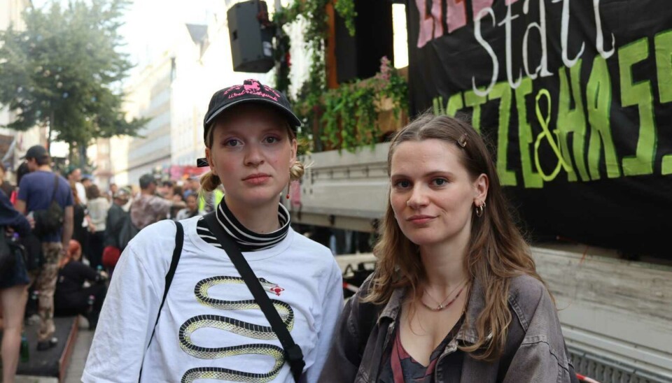 Zwei Personen stehen vor einem Protestbanner, dahinter versammelt sich eine Menschenmenge.