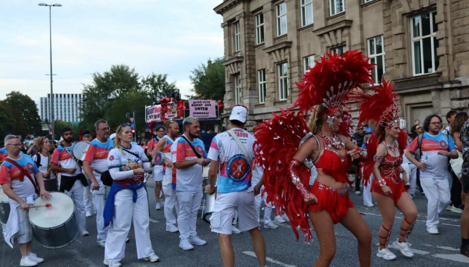 Tänzer in roten und weißen Kostümen führen eine Parade mit Trommeln durch eine belebte Innenstadtstraße an.