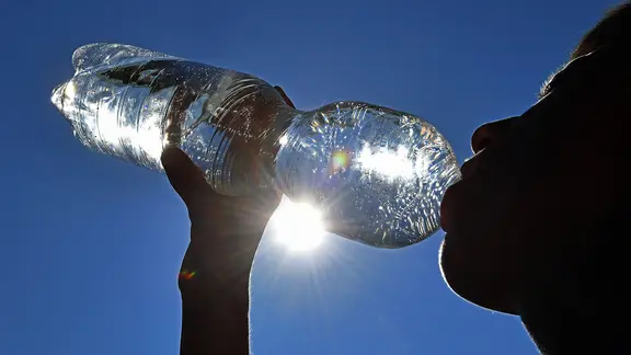 Eine Person trinkt Wasser aus einer Flasche, im Hintergrund Sonne und blauer Himmel | IMAGO Eine Person trinkt Wasser aus einer Flasche, im Hintergrund Sonne und blauer Himmel