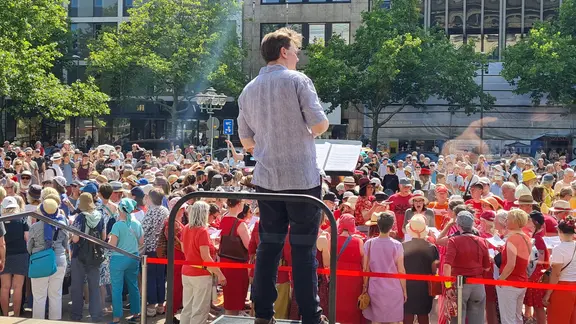 Ein Mann, von hinten zu sehen, steht auf einer Bühne. Vor ihm stehen viele Menschen. Er dirigiert ein Lied, das alles mitsingen. | NDR, Agnes Bührig Ein Mann, von hinten zu sehen, steht auf einer Bühne. Vor ihm stehen viele Menschen. Er dirigiert ein Lied, das alles mitsingen.