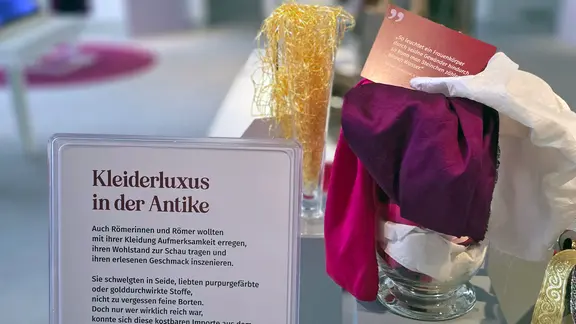 Gold, Seide und die Farbe der Purpurschnecke: Die, die es sich leisten konnten, stachen auch schon in der Antike mit ihrer Kleidung heraus. | NDR, Foto: Agnes Bührig Gefärbte Garne und Tücher stehen mit Erklärtexten versehen in einer Ausstellung.