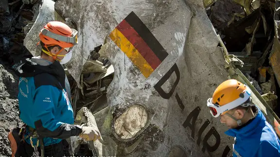 Zwei Bergungskräfte stehen vor einem Wrackteil eines Flugzeugs.