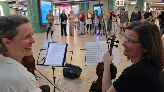 Zwei Musikerinnen sitzen mit ihren Instrumenten, einer Geige und einem Cello, vor Notenständern und lächeln sich gegenseitig an. Im Hintergrund stehen zahlreiche Menschen in einer Bahnhofshalle und beobachten das Duo. | NDR, Foto: Agnes Bührig Zwei Musikerinnen sitzen mit ihren Instrumenten, einer Geige und einem Cello, vor Notenständern und lächeln sich gegenseitig an. Im Hintergrund stehen zahlreiche Menschen in einer Bahnhofshalle und beobachten das Duo.