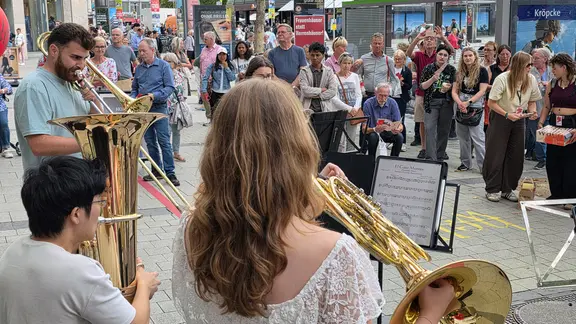 Auch für das Quintett Neues Haus Brass bleiben am Samstag viele Menschen, die in Hannovers Innenstadt unterwegs sind, stehen. | NDR, Foto: Agnes Bührig Drei Musiker spielen Blechblasinstrumente auf einer belebten Fußgängerzone, während eine große Gruppe von Zuschauern aufmerksam zuhört.