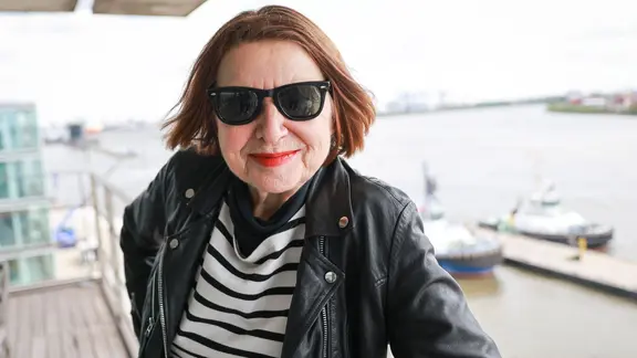 Inga Rumpf steht auf einer Terrasse mit Blick auf den Hamburger Hafen, trägt eine schwarze Sonnenbrille, eine Lederjacke und ein gestreiftes Shirt. | Christian Charisius/dpa +++ dpa-Bildfunk +++ Inga Rumpf steht auf einer Terrasse mit Blick auf den Hamburger Hafen, trägt eine schwarze Sonnenbrille, eine Lederjacke und ein gestreiftes Shirt.