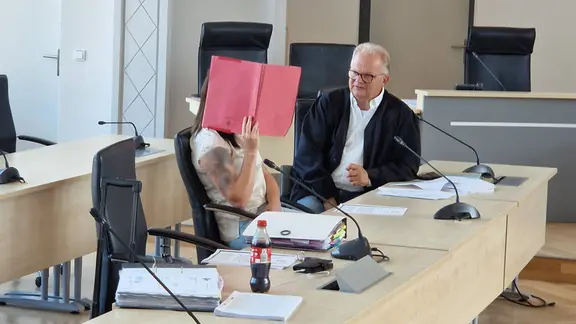 Gina H. sitzt neben ihrem Verteidiger und hält sich einen Aktendeckel vors Gesicht. | Andreas Frost, NDR Gina H. sitzt neben ihrem Verteidiger und hält sich einen Aktendeckel vors Gesicht.