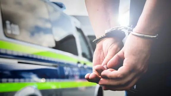 Symbolbild - Ein Mann wird mit Handschellen von der Polizei festgenommen. Im Hintergrund steht ein Streifenwagen der Polizei mit reflektierenden Markierungen. | IMAGO / Fotostand Symbolbild - Ein Mann wird mit Handschellen von der Polizei festgenommen. Im Hintergrund steht ein Streifenwagen der Polizei mit reflektierenden Markierungen.