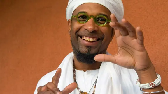 Ein wesentlicher Teil der Musik Mittel- und Südamerikas hat afrikanische Ursprünge - diese Erkenntnis lebt der kubanische Pianist Omar Sosa schon seit Langem. | Tom Ehrlich Omar Sosa lächelt und steht in weißer Kleidung und mit einer grünen Brille auf vor einer orangefarbenen Wand. Er hält die Hände in einer offenen, gestikulierenden Pose und trägt mehrere Halsketten und Armbänder.