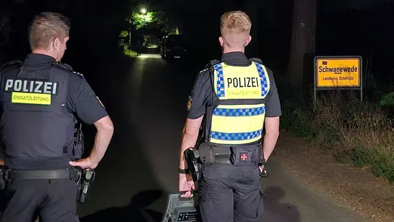 Zwei Polizisten stehen mit einem mobilen Scheinwerfer am Ortseingang von Schwanewede. Im Fall des getöteten Ehepaares in Bremen hat die Polizei den mutmaßlichen Täter festgenommen.