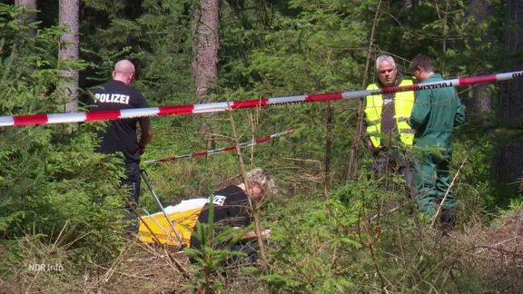 Vier Personen und ein Flugzeugteil hinter einer Polizeiabsperrung im Wald. | Screenshot Vier Personen und ein Flugzeugteil hinter einer Polizeiabsperrung im Wald.