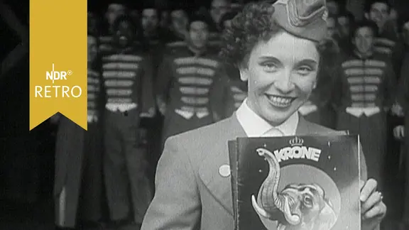 Ansagerin präsentiert Programmheft des Zirkus Krone vor Zirkusmitarbeitern in Uniformen (1956) | Screenshot Ansagerin präsentiert Programmheft des Zirkus Krone vor Zirkusmitarbeitern in Uniformen (1956)