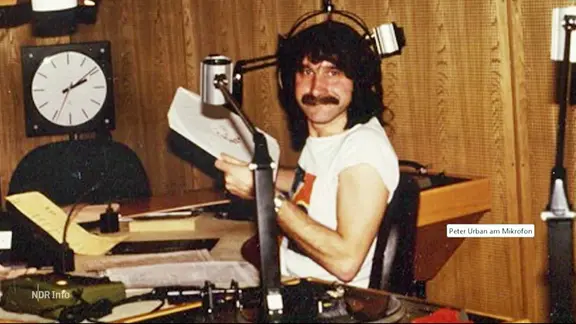 Peter Urban in jungen Jahren. Er sitzt in einem Studio, trägt lange Haare und einen Schnauzer.