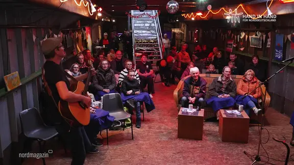 Menschen sitzten im Frachtraum eines Schiffes und singen gemeinsam. | Screenshot Menschen sitzten im Frachtraum eines Schiffes und singen gemeinsam.