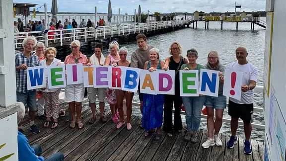 Die "Initiative Weiterbaden!" fordert den Erhalt des historischen Seebads. | Tina Loeffelbein Menschengruppe mit einem "Weiterbaden"-Schild am Seebad Düsternbrook.