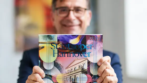 Intendant Bodo Busse lächelt und hält ein bunt gestaltetes Programmheft mit der Aufschrift "Staatsoper Hannover" in die Kamera. | Clemens Heidrich Intendant Bodo Busse lächelt und hält ein bunt gestaltetes Programmheft mit der Aufschrift "Staatsoper Hannover" in die Kamera.