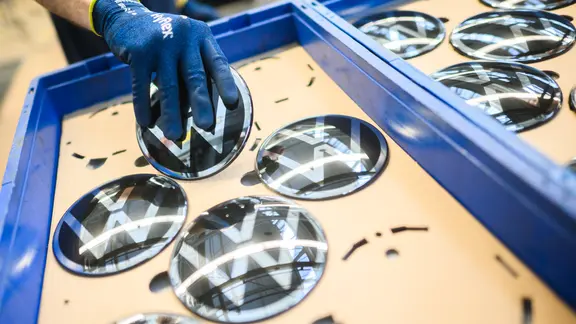 Ein Mitarbeiter hält im VW Stammwerk ein VW Logo für den Einbau in einen Neuwagen. | dpa-Bildfunk, Julian Stratenschulte Ein Mitarbeiter hält im VW Stammwerk ein VW Logo für den Einbau in einen Neuwagen.