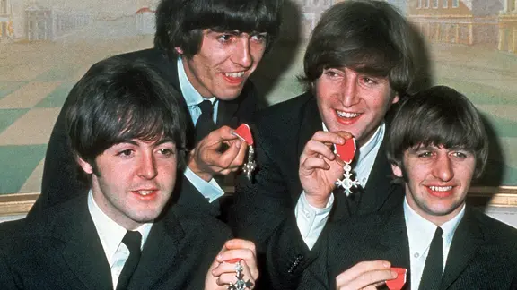 Mit der Pilzkopf-Frisur sind die Beatles um John Lennon berühmt geworden. | dpa/picture-alliance Paul McCartney, George Harrison, John Lennon und Ringo Starr mit den Orden "Member of the Order of the British Empire" (Aufnahme vom 26. Oktober 1965)