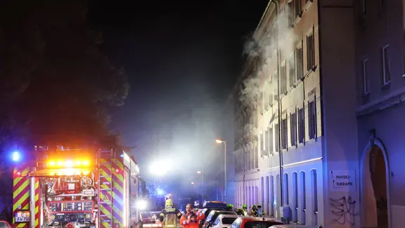 Ein Rettungswagen und Feuerwehrleute stehen vor einem Wohnhaus, aus dem Rauch quillt. 