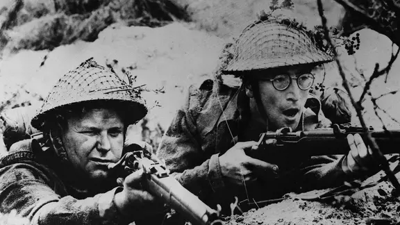 Das Filmmuseum Bendestorf widmet den Dreharbeiten zu "Wie ich den Krieg gewann" einen eigenen Ausstellungsraum. Auch Vorführungen des Films fanden hier schon statt. | Imago / KPA Zwei Männer (rechts John Lennon als Gefreiter Gripweed) liegen auf dem Bauch und zielen mit Gewehren nach vorn (Filmaufnahme von 1966)