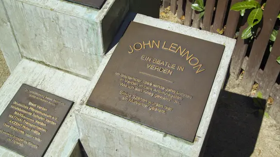 Im niedersächsischen Verden erinnert heute ein Denkmal an die Dreharbeiten mit John Lennon. | IMAGO / CHROMORANGE Eine Gedenktafel auf einem Betonblick mit der Aufschrift: "John Lennon. Ein Beatle in Verden"
