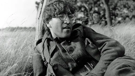 John Lennon lehnt mit kurzen Haaren, Nickelbrille und Soldatenuniform an einem Baumstamm. | IMAGO / Keystone Press John Lennon lehnt mit kurzen Haaren, Nickelbrille und Soldatenuniform an einem Baumstamm.
