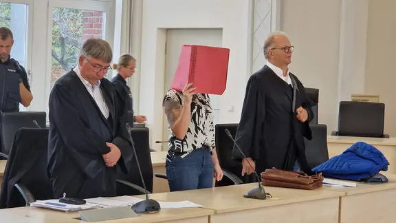 Die Angeklagte Gina H. hält sich einen Aktenordner vor das Gesicht und steht zwischen ihren Anwälten. | Andreas Frost, NDR Die Angeklagte Gina H. hält sich einen Aktenordner vor das Gesicht und steht zwischen ihren Anwälten.