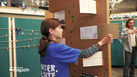Ein Kind mit langen Haaren öffnet eine Tür an einem Haus aus Pappe. | Screenshot Ein Kind mit langen Haaren öffnet eine Tür an einem Haus aus Pappe.
