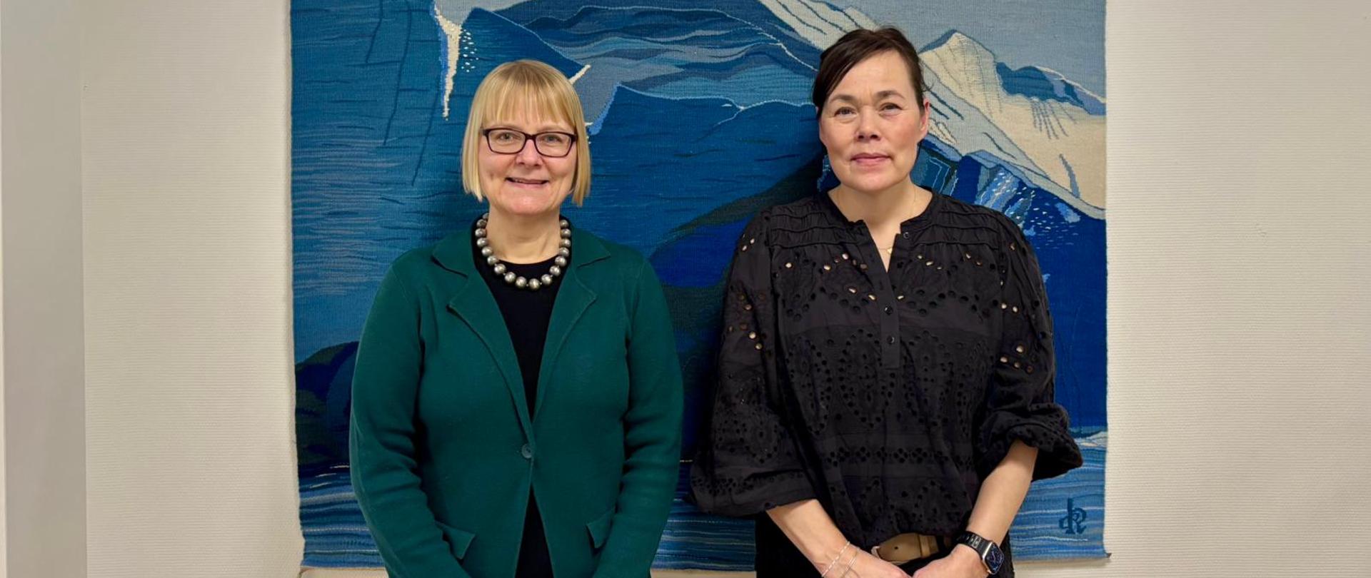 Women posing, on the left, Ambassador Ewa Dębska wearing dark green blazer; on the right, Minister of Foreign Affairs and Research Vivan Motzfeldt, wearing black blouse, standing together against a textile depicting a mountainous landscape.