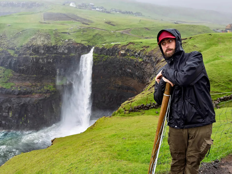 Person in rain gear poses by a waterfall on a lush green landscape, with mist in the background