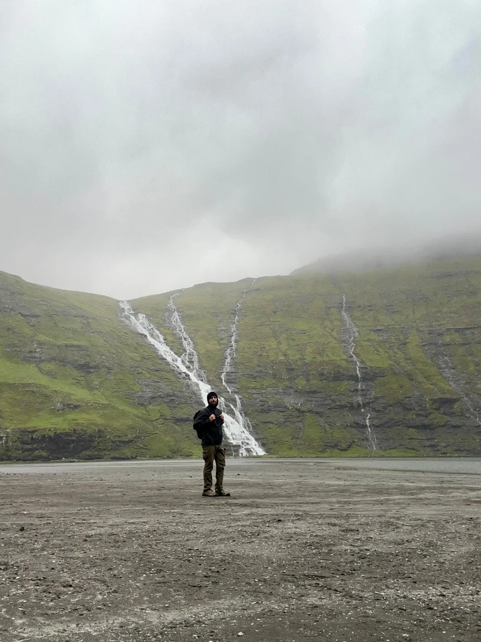Person standing on a misty plain with a backdrop of cascading waterfalls and grassy hills, creating a serene, natural setting