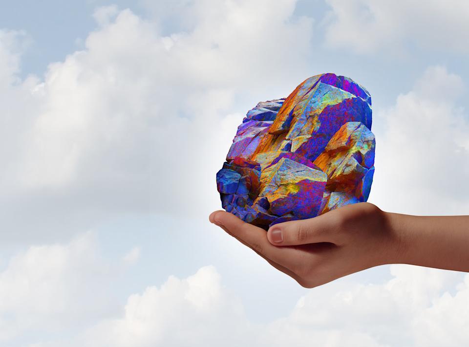 Image shows a hand holding up a colorful rock, meant to represent rare-earth elements, against a cloudy sky in the background.