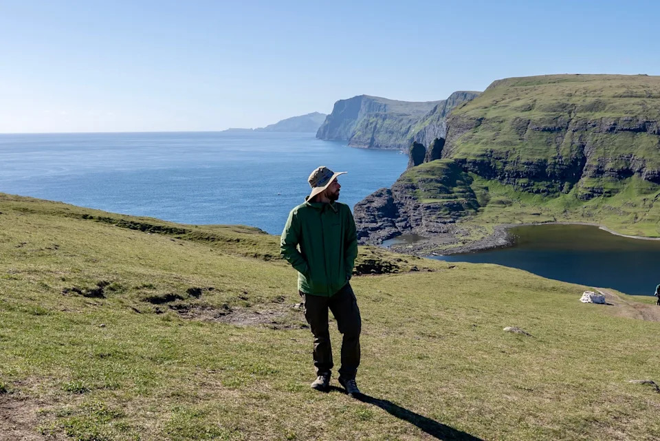 A person in outdoor attire stands on a grassy cliff overlooking the ocean and rugged coastline, capturing the essence of a scenic travel destination