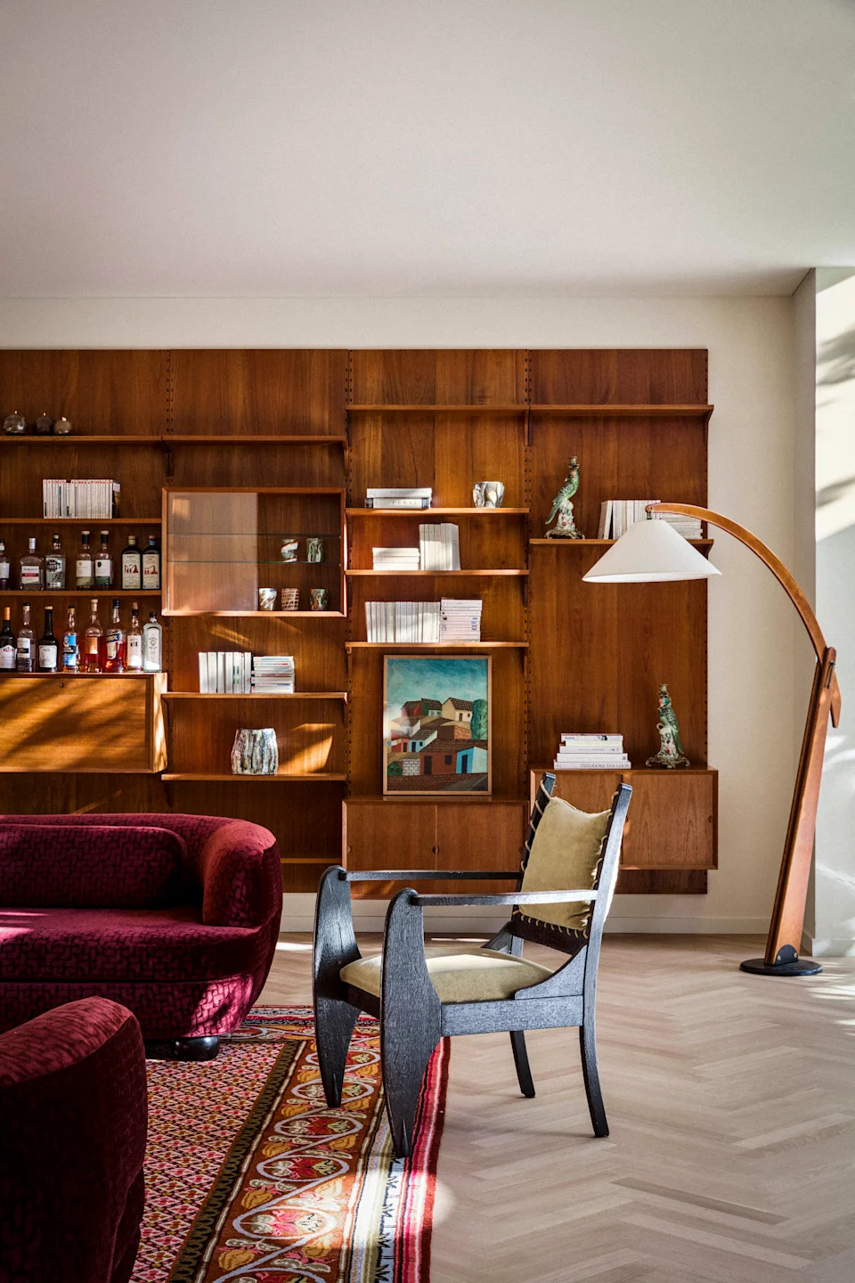 A living room with a red printed rug, a deep toned velvet sofa, an armchair, a standing lamp and a bookshelf